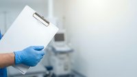 Doctor or healthcare professional wearing blue latex gloves holding blank clipboard in medical clinic or hospital setting with blurred equipment background.