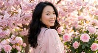 Young Asian woman smiling joyfully among blooming pink cherry blossoms and roses in spring garden setting for lifestyle and beauty content.