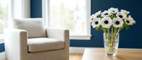 White anemone flowers in a crystal vase on a wooden table, with a cozy armchair and bright window in a modern living room.
