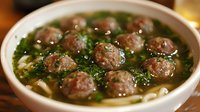 Homemade meatball soup with noodles in clear broth, garnished with fresh herbs and green onions, served in white ceramic bowl. Traditional comfort food.