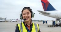 Young Asian female airport ground crew worker wearing headset and high visibility vest smiling at camera with commercial aircraft in background at busy airport tarmac.