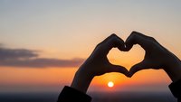 Hands forming heart shape silhouette against golden sunset sky. Romantic love symbol gesture at twilight for Valentine's Day greeting cards and wedding designs.