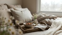Cozy bedroom morning scene with breakfast tray on unmade bed featuring linen bedding, natural light streaming through window creating peaceful atmosphere.