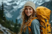 Young Caucasian woman hiker with long blonde hair wearing beanie and yellow backpack smiles while exploring mountain wilderness against backdrop of misty peaks and evergreens.