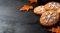 Homemade ginger cookies decorated with white snowflake icing on dark stone background with orange autumn maple leaves. Winter holiday baking concept.