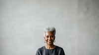 Senior woman with short white hair and dark skin, wearing casual dark clothing, smiling confidently against neutral gray background. Portrait suitable for diversity, aging, wellness and lifestyle content.