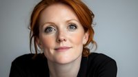 Young redhead Caucasian woman with natural makeup and updo hairstyle wearing black top smiling at camera against light gray background, soft lighting portrait.