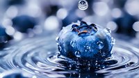 Fresh blueberry with water drops splashing in clear water creating concentric ripples, captured in macro photography with bokeh background in blue tones.