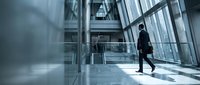 Professional businessman walking through modern glass office building corridor with natural light streaming through windows creating dramatic shadows.