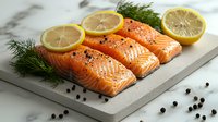 Fresh raw salmon fillets seasoned with black pepper and lemon slices on white marble board, garnished with dill sprigs for gourmet cooking preparation.