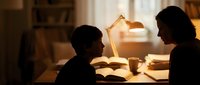 Mother and child reading together at desk with warm lamp light creating cozy evening study atmosphere in home interior for family education concepts.