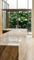 Modern marble kitchen island with natural stone countertop in contemporary home interior featuring floor-to-ceiling windows and lush tropical garden view.