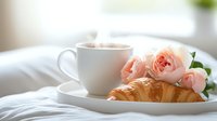 Romantic breakfast setting with steaming coffee cup, fresh buttery croissant and delicate pink garden roses on white linens. Morning light creates soft ambiance.