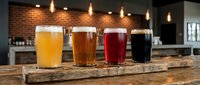 Four craft beer glasses with different colored brews displayed on rustic wooden board in modern brewery or pub setting with warm lighting and brick wall background.