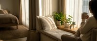 Young woman with dark hair and light skin tone, wearing casual beige clothing, sitting by a sunlit window in a modern bedroom. Natural light streams through sheer curtains, illuminating potted green plants on the windowsill. Cozy interior design with neutral tones, comfortable seating, and minimalist decor. Suitable for lifestyle, wellness, home design, and relaxation concepts.