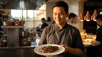 Male chef of Asian descent wearing dark chef's attire presenting plated meat dish in busy restaurant kitchen with staff and pendant lighting in background.