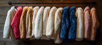 Colorful faux fur coats hanging on clothing rack against wooden wall background. Fashion retail display of trendy winter outerwear in white, pink, blue and beige tones.