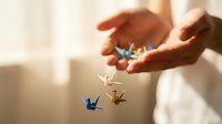 Hands holding and releasing origami paper cranes in blue and gold, symbolizing hope, wishes, and new beginnings for inspirational and wellness content.