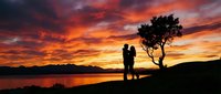 Silhouetted couple standing together by a lake at sunset with dramatic orange and red sky, golden hour landscape for romantic and travel concepts.