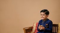 Young boy with dark hair and South Asian ethnicity wearing traditional blue kurta with gold embroidery, seated on wooden chair against neutral beige background, smiling while gesturing with hand.