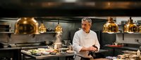 Confident middle-aged Caucasian male chef in white uniform standing in professional restaurant kitchen with golden pendant lights and steaming pots.