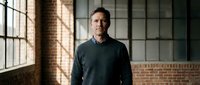 Middle-aged Caucasian businessman in dark sweater standing confidently in modern loft office with brick walls and large windows for corporate presentations.