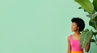 Young African American woman wearing bright pink sleeveless top standing against mint green background with tropical banana plant leaves, looking upward with positive expression for lifestyle and wellness content.
