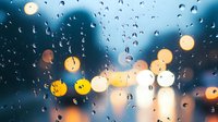Raindrops on window glass create bokeh effect with blurred city lights in background during evening rain, moody atmospheric urban scene with water droplets.