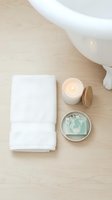 Spa and wellness setup with folded white towel, lit candle with wooden lid, soap bar, and ceramic bowl on light wood surface for relaxation and self-care.