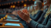 Young woman applauding during conference presentation, with blurred audience and glowing tablets in dark auditorium creating atmospheric bokeh lighting effect.