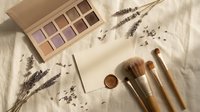 Natural beauty cosmetics flat lay with eyeshadow palette, makeup brushes and dried lavender flowers on neutral linen background for skincare branding.