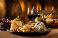 Gourmet cheese platter with blue cheese, aged cheddar, fresh grapes, and red wine served in rustic setting with warm ambient lighting and wooden barrel background.