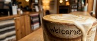 Rustic ceramic mug with latte art and Welcome inscription on wooden table in cozy cafe interior with warm bokeh background.