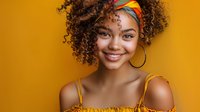 Young African American woman with natural curly hair wearing yellow top and colorful headband smiling warmly against vibrant orange background. Positive lifestyle portrait.