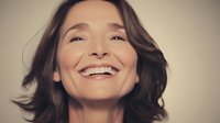Mature Caucasian woman with natural makeup and brown wavy hair showing genuine happy smile against beige background, expressing joy and confidence in lifestyle portrait.