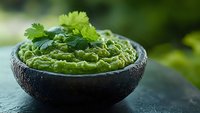 Fresh guacamole dip garnished with cilantro leaves served in dark stone bowl against blurred green background, traditional Mexican appetizer for parties.