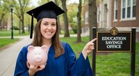 Young female graduate in blue cap and gown holding pink piggy bank on college campus, celebrating education savings and financial planning for students.