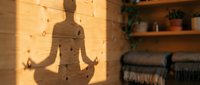 Silhouette of a person in lotus meditation pose cast on wooden wall in warm sunlight, with shelves and blankets visible, conveying mindfulness and relaxation for wellness content.