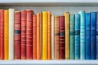Colorful vintage hardcover books arranged in rainbow order on white bookshelf, from warm reds and oranges to cool blues, showing spines with gold lettering.