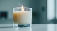 Glowing white scented candle in clear glass holder on blurred teal background, creating cozy atmosphere with soft smoke trail from burning flame.