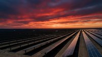Rows of solar panels stretch across agricultural land beneath a dramatic sunset sky with vibrant orange and red clouds, representing renewable energy and sustainable power generation technology.