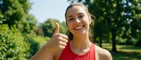 Happy young Caucasian woman in red sports top giving thumbs up gesture outdoors in sunny park after workout, smiling at camera with positive attitude.