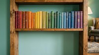 Colorful rainbow books arranged on wooden shelf creating vibrant spectrum from red to purple against green wall in cozy reading room interior.