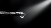 Single water droplet hanging from textured surface edge in dramatic black and white macro photography with soft lighting and dark background.