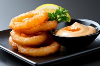 Crispy golden onion rings stacked on black plate with creamy dipping sauce, garnished with fresh parsley and lemon slice, close up view on dark background.