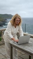 Middle-aged woman with shoulder-length blonde hair and glasses working on laptop at wooden table on coastal terrace overlooking ocean and green cliffs. Remote work concept.