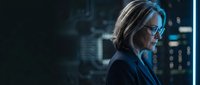 Mature woman with shoulder-length blonde hair and glasses wearing dark business suit, profile view against illuminated cityscape backdrop with blue neon lighting, corporate professional environment.