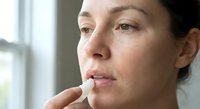 Young woman applying lip balm stick to lips for skincare routine and moisturizing treatment in natural daylight by window.