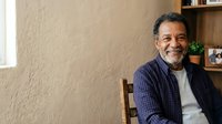Middle-aged man with dark hair and beard, South Asian ethnicity, wearing navy blue checkered shirt over white undershirt, sitting indoors by window with wooden shelving behind him, smiling at camera.