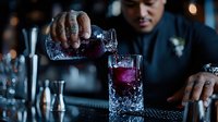 Asian male bartender with tattoos pouring purple craft cocktail into crystal glass at upscale bar. Professional mixology and modern drink preparation in dark ambient setting.